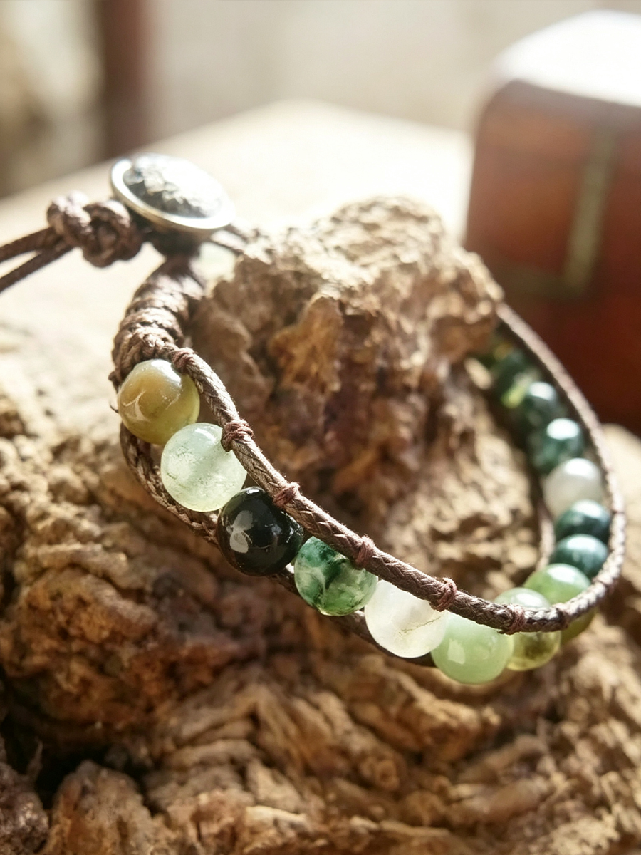 A close-up of a handmade wrap bracelet with brown cord, green and white translucent beads, and a silver-toned charm, displayed on a textured piece of wood.
