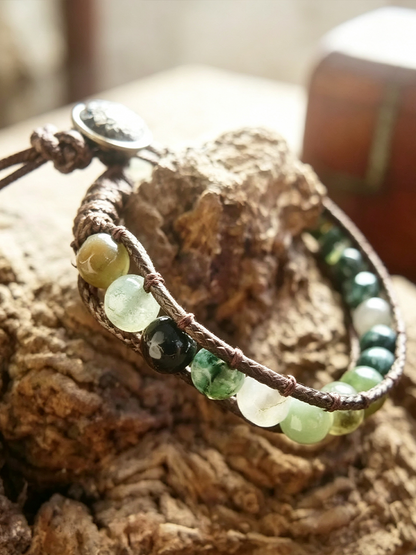 A close-up of a handmade wrap bracelet with brown cord, green and white translucent beads, and a silver-toned charm, displayed on a textured piece of wood.