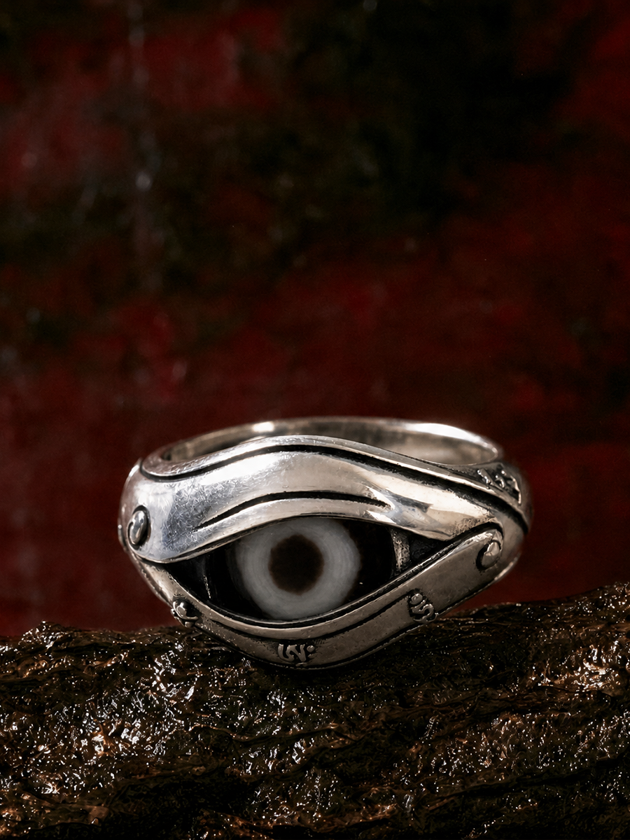 A sterling silver evil eye ring with a detailed black and white eye motif, resting on a textured dark brown surface against a dark red background.