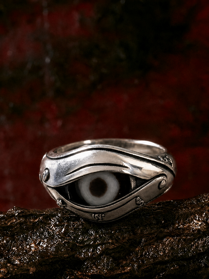 A sterling silver evil eye ring with a detailed black and white eye motif, resting on a textured dark brown surface against a dark red background.