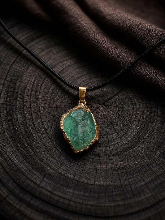 A raw green stone pendant with a gold-toned border, suspended on a black cord necklace, displayed on a dark wood slice background.