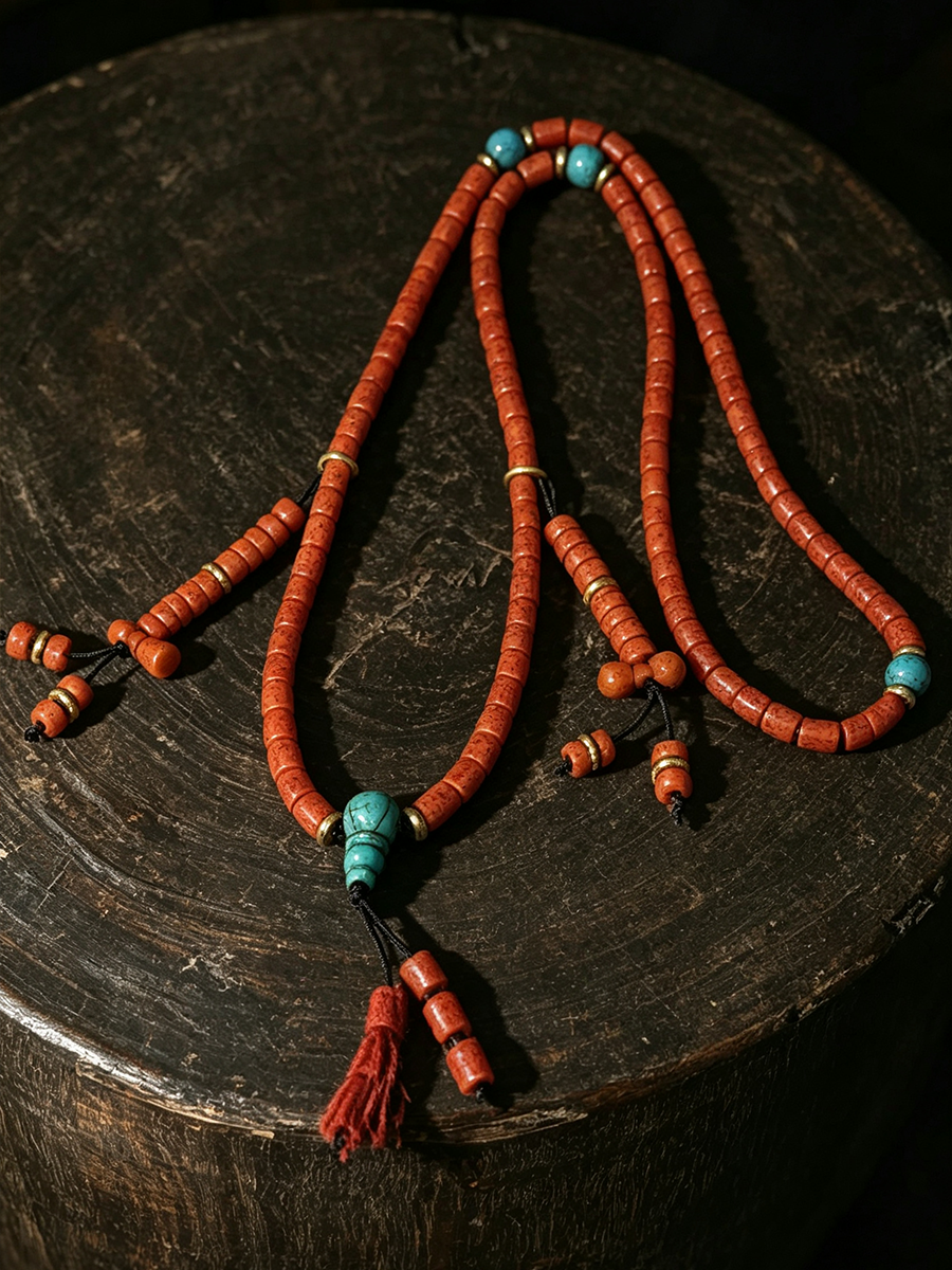 A beaded necklace with terracotta-colored cylindrical beads, turquoise blue accent beads, and a red tassel, displayed on a dark wooden surface.