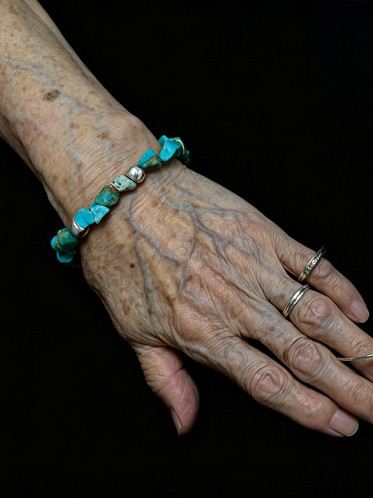 An elderly hand and wrist wearing a beaded bracelet made of turquoise-blue stones and silver-toned beads, shown against a solid black background.