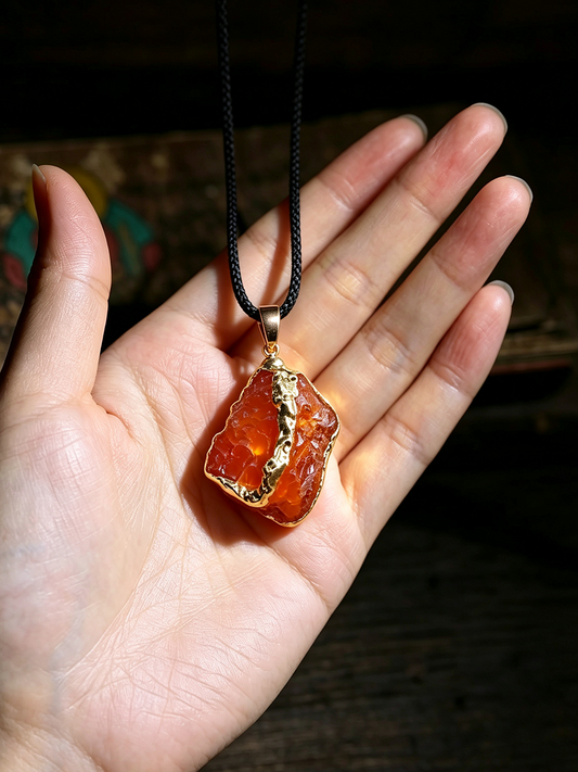 A raw orange stone pendant with gold trim hanging from a black cord, held in a person's hand against a dark background.