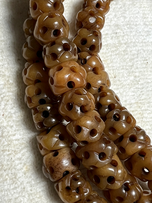 Close-up of carved brown beads with multiple drilled holes, arranged in strands on a light textured fabric background.