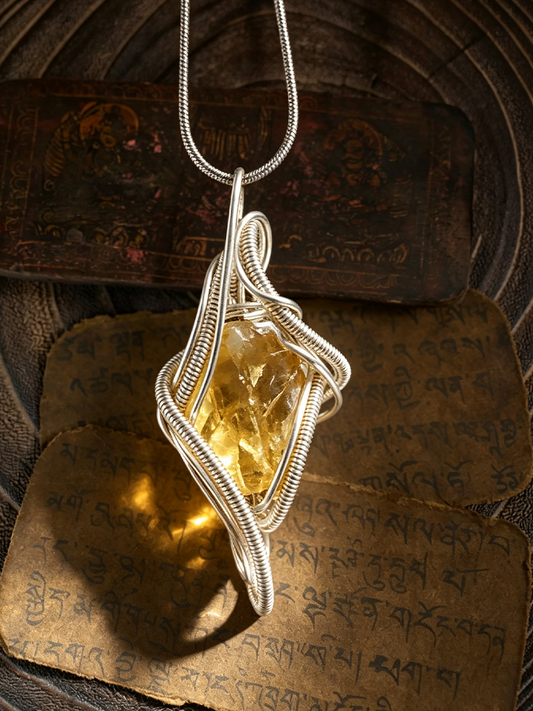 A silver wire-wrapped pendant necklace featuring a faceted yellow crystal, displayed on a dark textured surface with Tibetan script cards in the background.