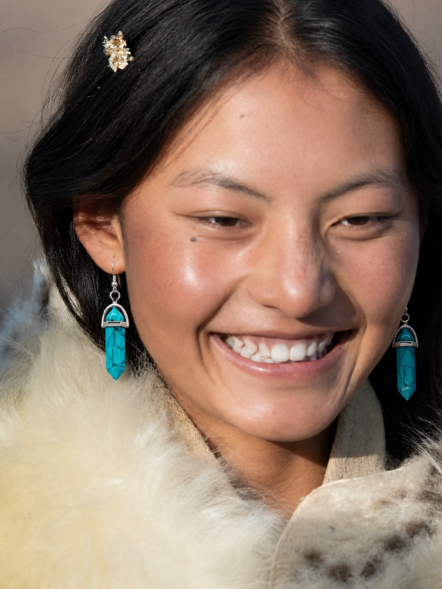 A close-up portrait of a smiling woman wearing turquoise drop earrings with silver-toned metal tops and dangling chains, along with a cream fur collar and a small gold hair accessory.