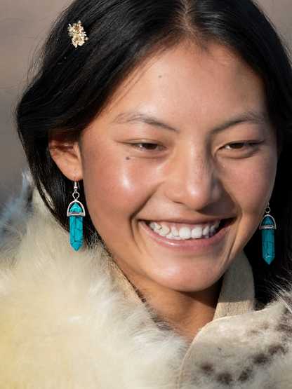 A close-up portrait of a smiling woman wearing turquoise drop earrings with silver-toned metal tops and dangling chains, along with a cream fur collar and a small gold hair accessory.