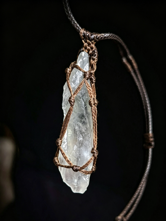 A raw, clear quartz crystal pendant wrapped in a rustic copper wire mesh, suspended from a dark brown adjustable cord necklace against a solid black background.