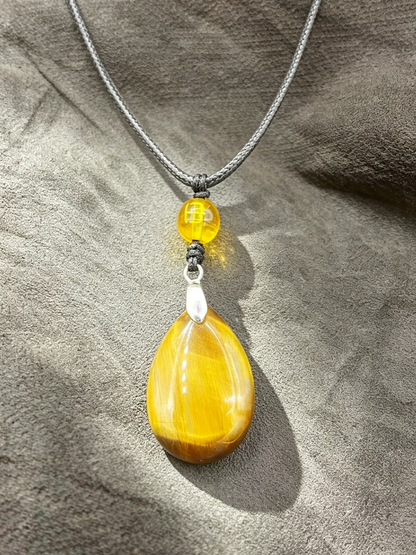 A yellow teardrop-shaped stone pendant necklace with a small yellow bead and silver spacer on a gray textured fabric background.