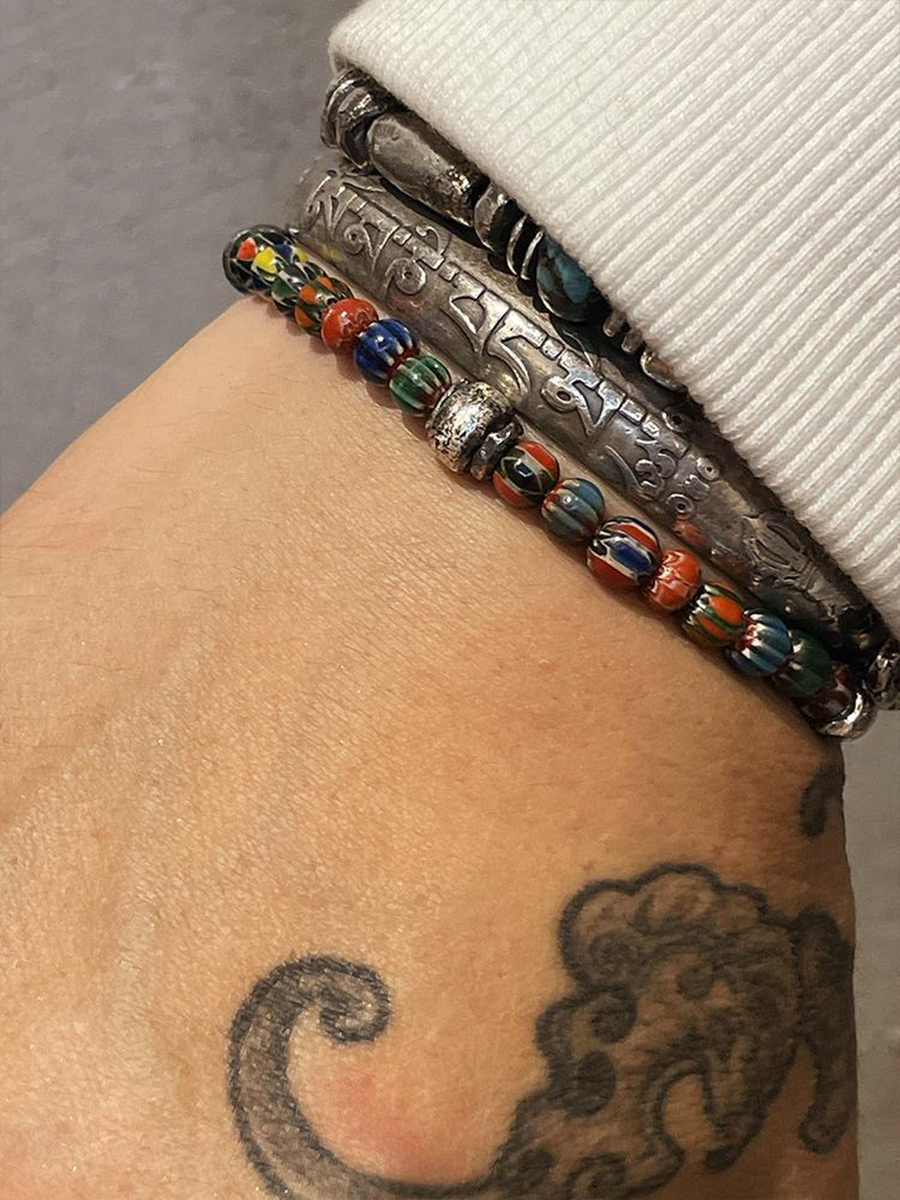 A close-up of a person's wrist wearing multiple beaded bracelets, including a silver engraved bracelet and a colorful beaded bracelet with red, blue, green, and yellow beads, next to a white ribbed cuff.