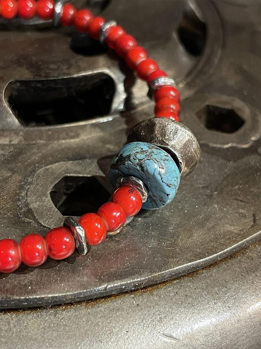 A close-up of a beaded necklace featuring bright red round beads, silver-toned spacers, and a large central turquoise-blue bead with a rustic silver cap, resting on a dark textured metal surface.