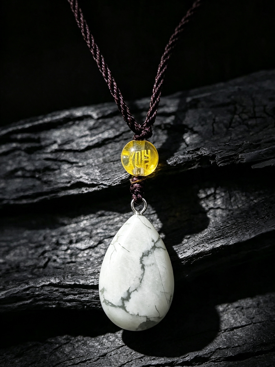 A teardrop-shaped white stone pendant with gray veining, suspended from a dark brown braided cord and a small round yellow bead, displayed on a dark textured wooden surface.