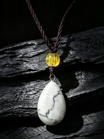 A teardrop-shaped white stone pendant with gray veining, suspended from a dark brown braided cord and a small round yellow bead, displayed on a dark textured wooden surface.