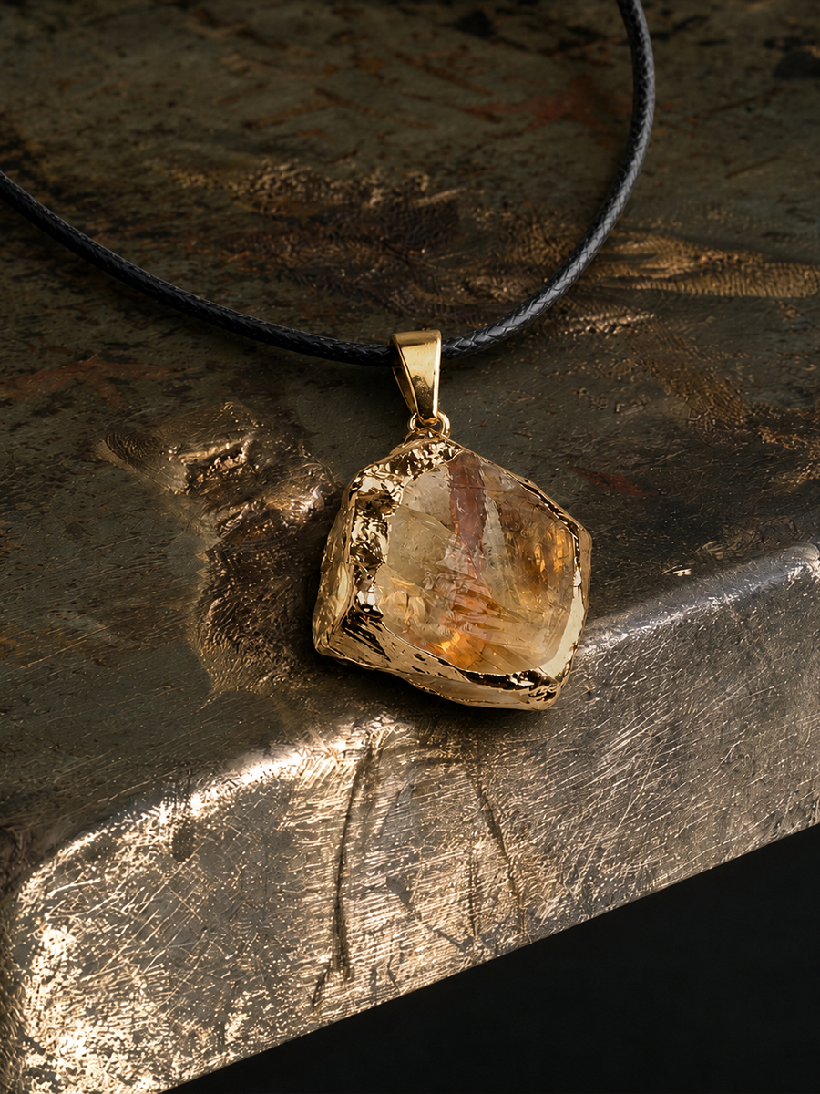 A raw, rough-cut amber and clear crystal pendant on a gold bail, suspended from a black cord, displayed on a textured metallic surface.