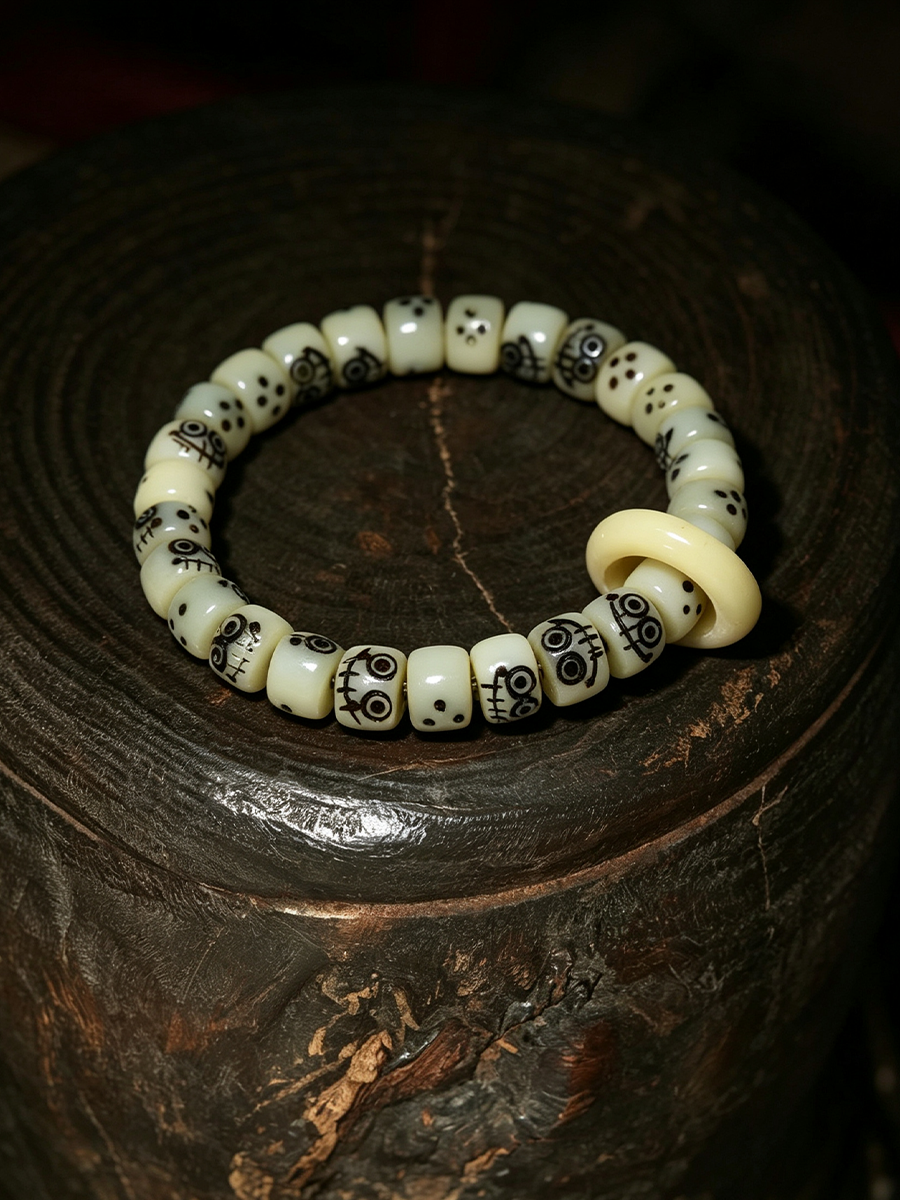 A beaded stretch bracelet made of cream-colored round beads with black printed patterns, featuring a larger flat cream-colored accent bead, displayed on a dark wooden surface.