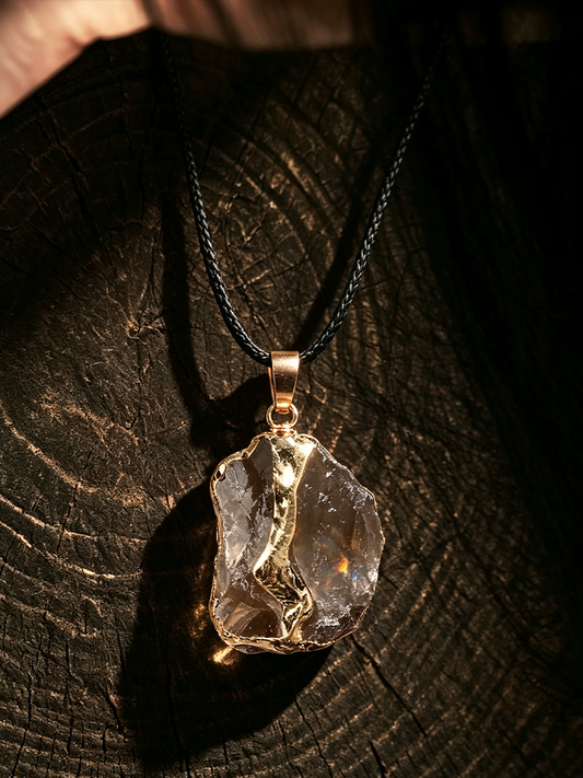 A raw, irregular clear gemstone pendant necklace with a gold-toned bezel and black cord, displayed on a dark textured wood surface.