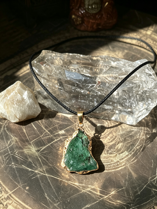 A raw green stone pendant necklace with a gold-toned bezel and black cord, displayed on a clear crystal cluster over a metallic surface.