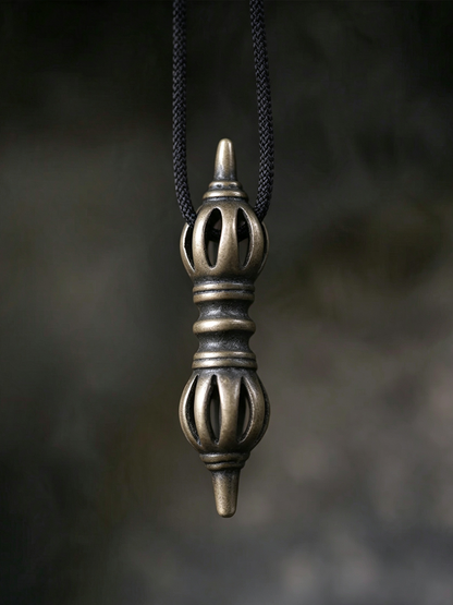 A bronze-colored metal pendant with a textured, ornate design hangs from a black braided cord against a dark, blurred background.