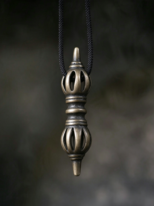 A bronze-colored metal pendant with a textured, ornate design hangs from a black braided cord against a dark, blurred background.