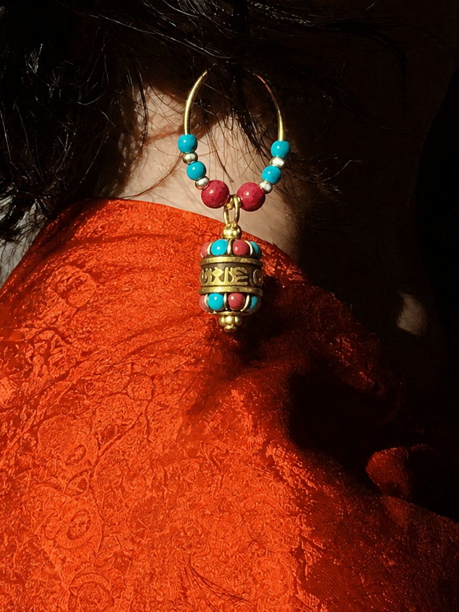 A close-up of a person wearing a large gold hoop earring with turquoise and red beads and a dangling ornate gold bell pendant, against dark hair and a bright orange textured garment.