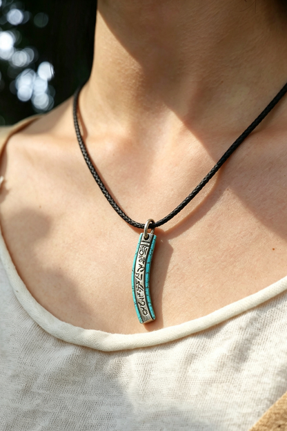 A close-up lifestyle shot of a person wearing a black braided cord necklace with a vertical turquoise and silver pendant.