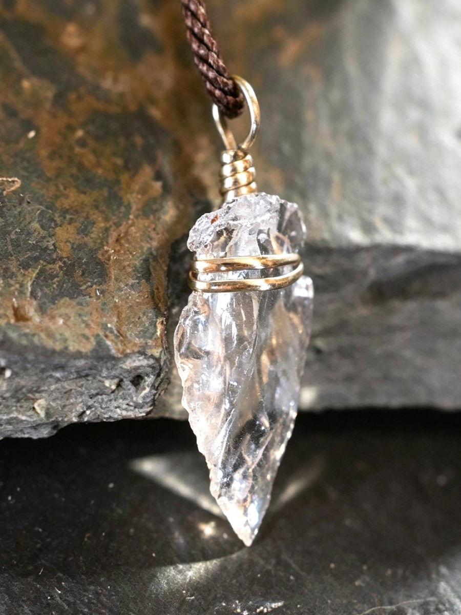 A clear crystal point pendant wrapped with gold wire, hanging from a dark brown cord, set against a dark textured rock background.