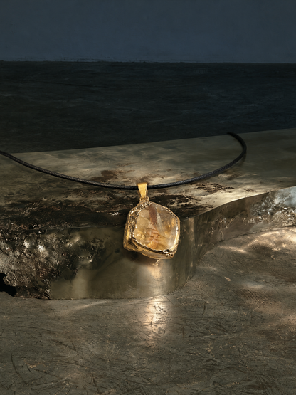 A gold pendant necklace with a rough, irregular-cut amber or honey-colored stone, resting on a dark concrete surface with a dark, moody background.