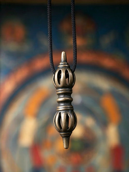 A metallic pendant necklace with a dark bronze or antique brass finish, suspended on a black cord against a blurred colorful background.
