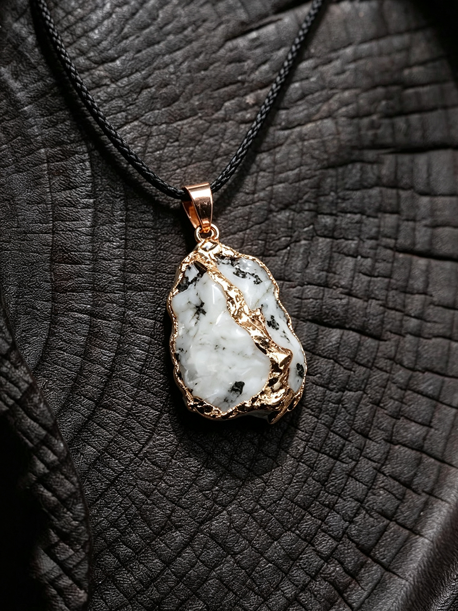 A raw white stone pendant with black inclusions, set in a rose gold bezel and hanging from a black cord, displayed on a textured black leather background.