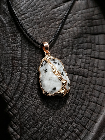 A raw white stone pendant with black inclusions, set in a rose gold bezel and hanging from a black cord, displayed on a textured black leather background.
