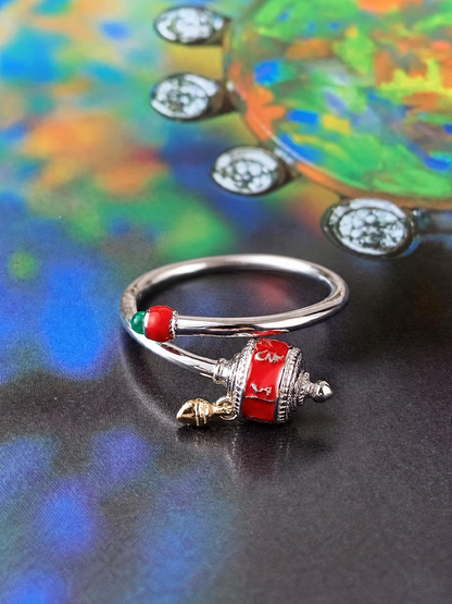 A silver-toned ring with a decorative red and green bead detail and a small gold-toned bell charm, photographed on a dark reflective surface with a colorful blurred background.