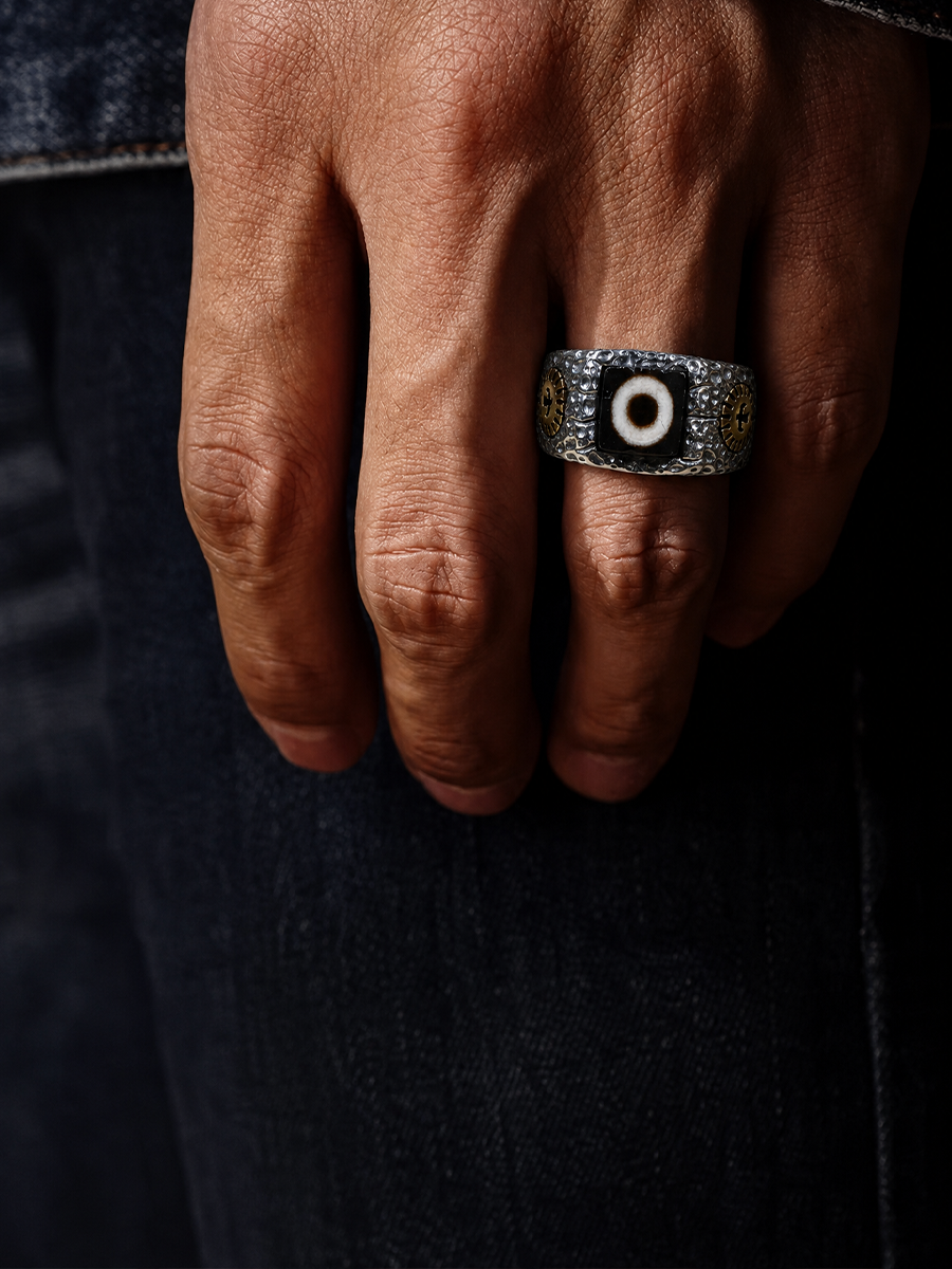 A close-up of a hand wearing a silver-toned signet ring with an evil eye motif, set against dark denim jeans.