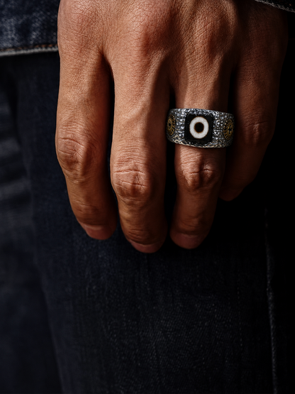 A close-up of a hand wearing a silver-toned signet ring with an evil eye motif, set against dark denim jeans.