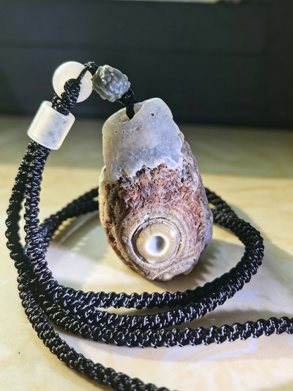 A shell pendant necklace featuring a natural oyster shell with a prominent eye motif, threaded onto a thick black braided cord with white and gray decorative beads.