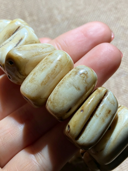 A close-up of a hand holding a bracelet made of large, polished, ribbed bone or ivory beads with natural brown and cream marbling.