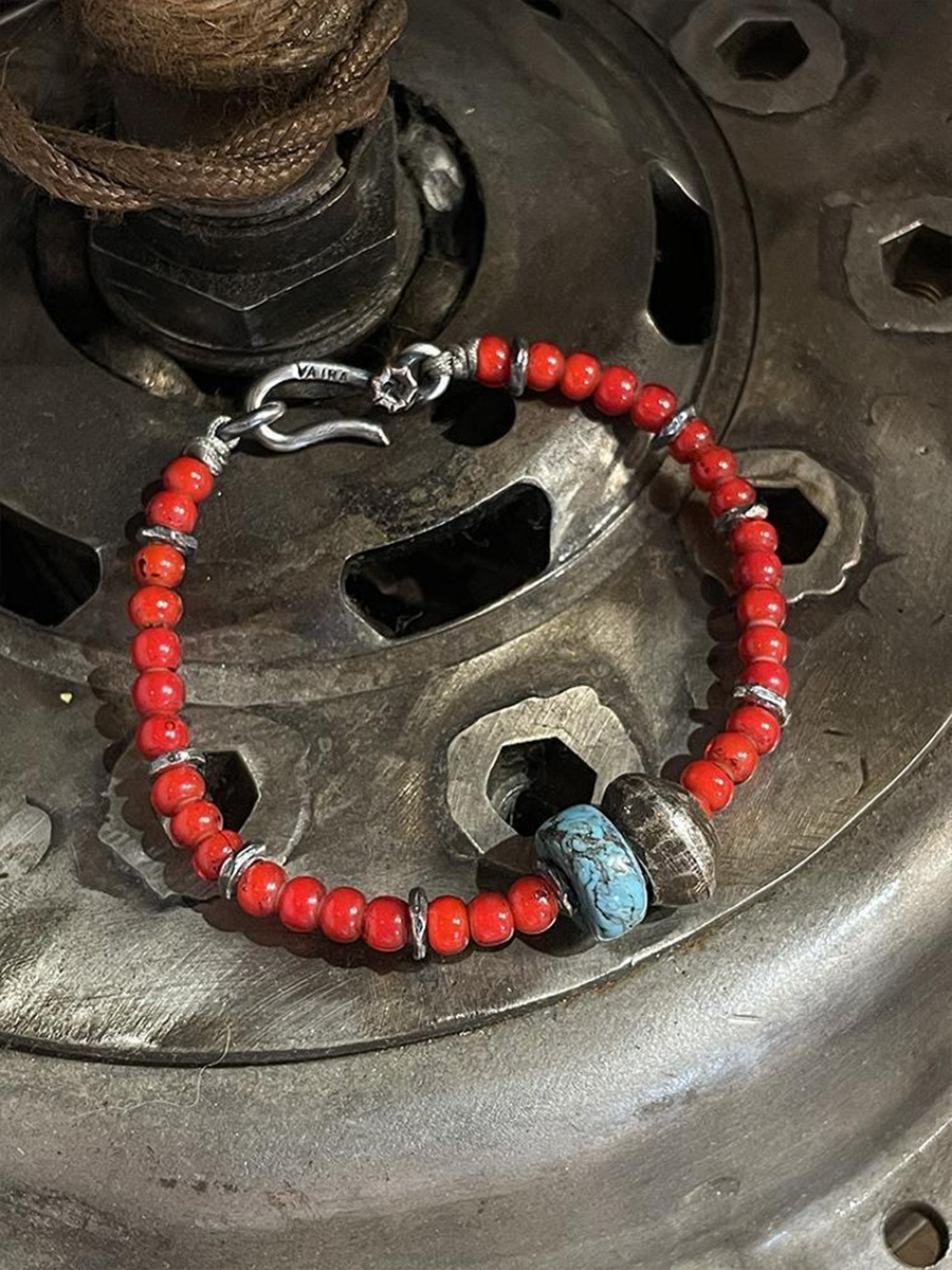 A beaded bracelet with small red-orange beads, silver-toned spacer rings, a silver hook clasp, and two larger accent beads including one turquoise-blue marbled bead, displayed on a metallic surface.