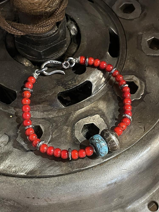 A beaded bracelet with small red-orange beads, silver-toned spacer rings, a silver hook clasp, and two larger accent beads including one turquoise-blue marbled bead, displayed on a metallic surface.