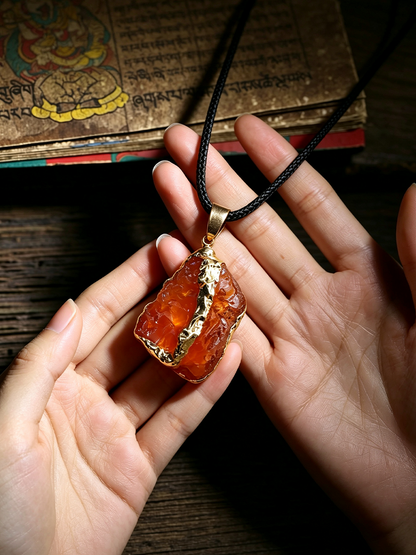 A person's hands holding a raw orange stone pendant necklace with gold trim and a black cord, set against a dark wooden background with an antique book.