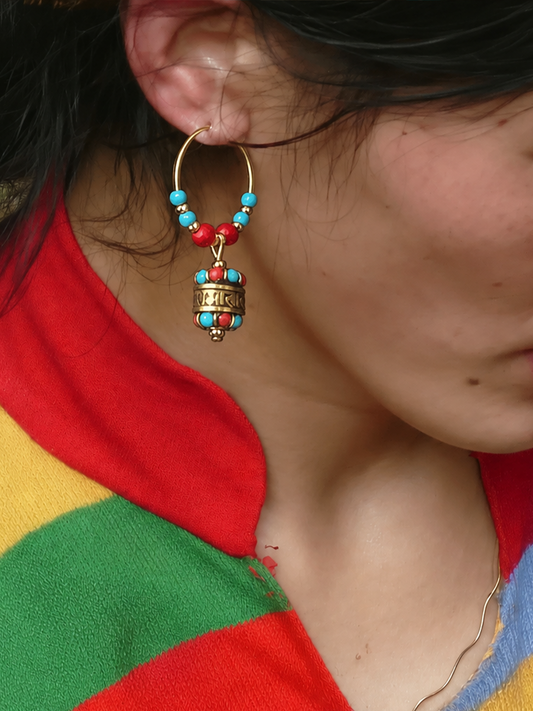 A close-up of a person wearing a gold hoop earring with turquoise and red beads and a dangling cylindrical charm, paired with a colorful striped sweater.