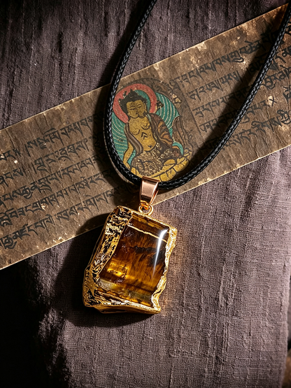 A raw tiger's eye pendant necklace with a gold-toned bezel and black cord, displayed on a textured brown linen fabric alongside a decorative printed paper strip.