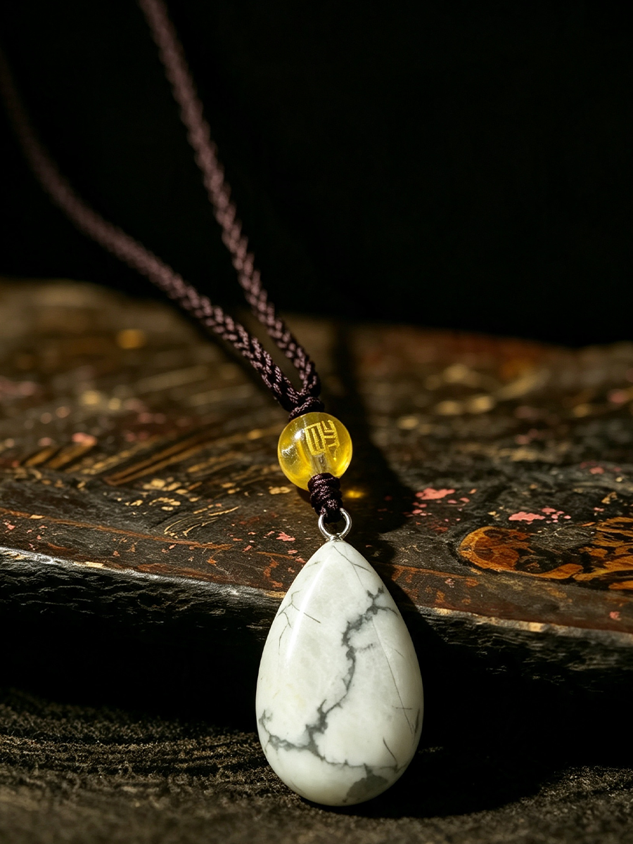 A teardrop-shaped white marble pendant necklace with dark veining, suspended from a dark brown braided cord with a small yellow bead, resting on a textured wooden surface against a dark background.