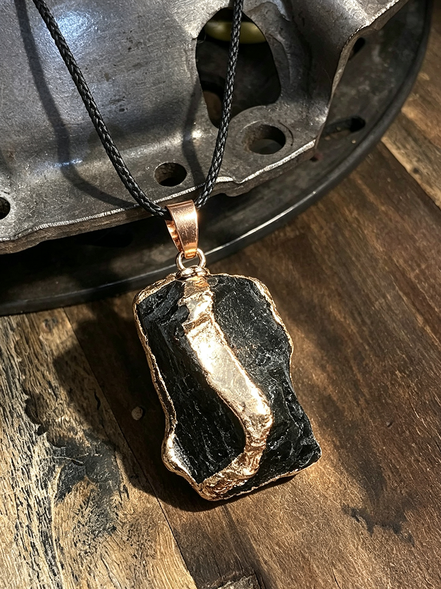 A handmade pendant necklace featuring a rough, textured black stone with a polished rose gold inlay, suspended on a black cord and resting on a rustic wooden surface next to a vintage metal wheel.