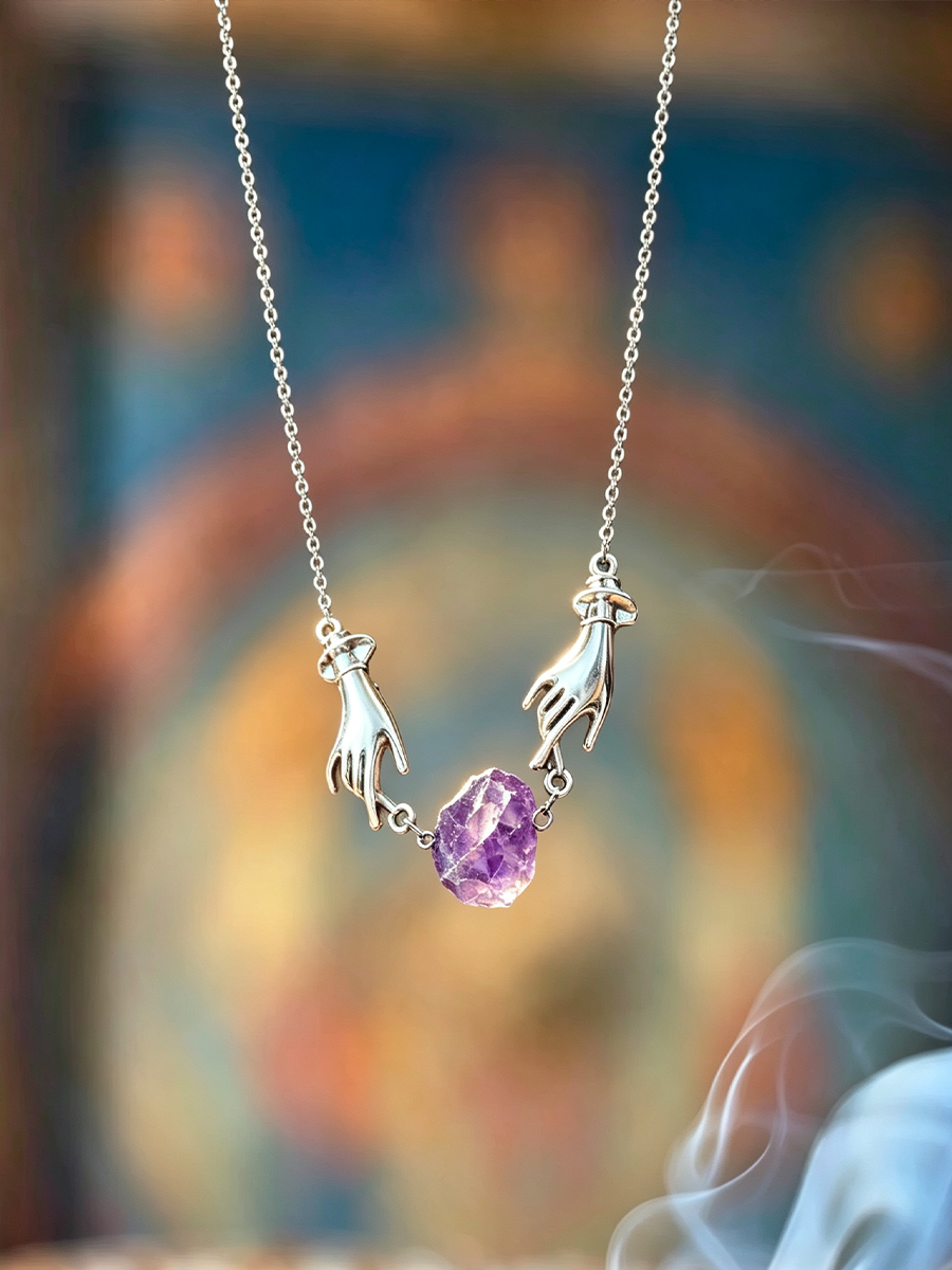 A silver necklace featuring two hands reaching toward a faceted purple amethyst stone, suspended from a delicate silver chain against a softly blurred, colorful background with wisps of smoke.