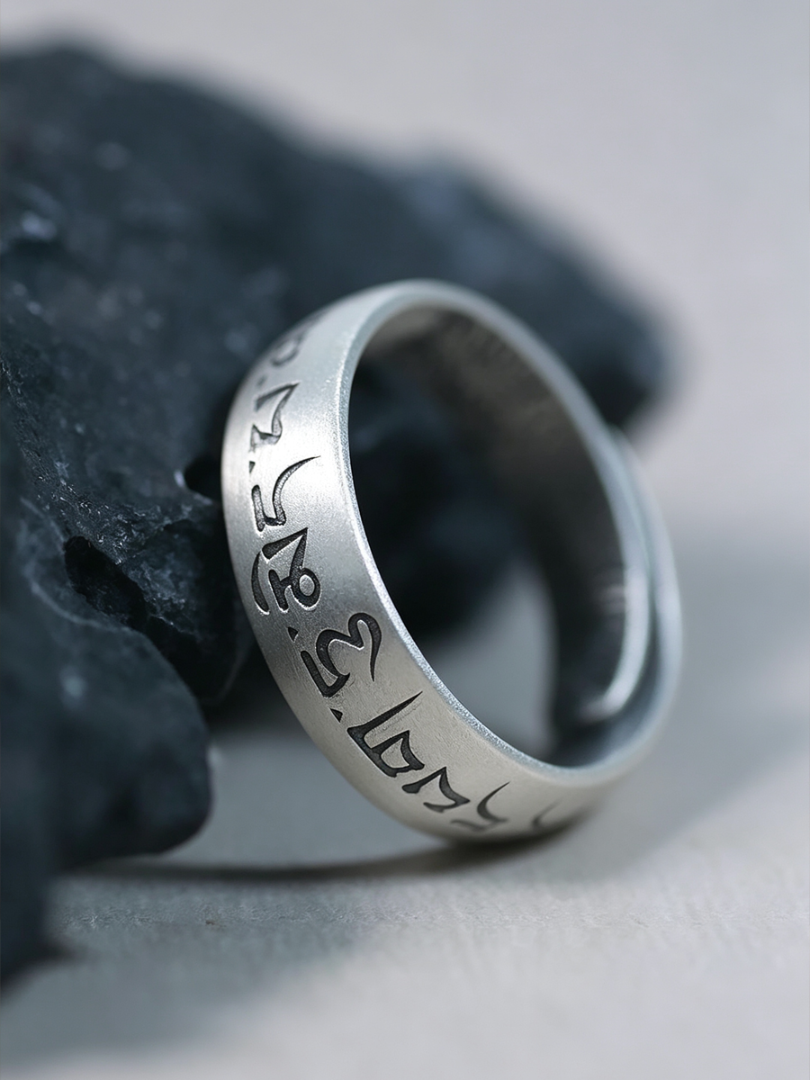 A silver-toned metal ring engraved with Tibetan script, resting on a dark textured stone against a light gray background.