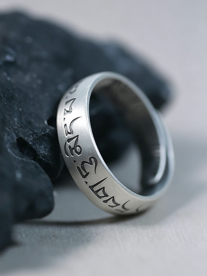 A silver-toned metal ring engraved with Tibetan script, resting on a dark textured stone against a light gray background.