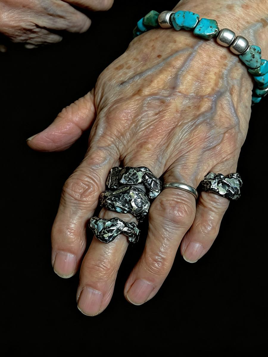 An elderly hand wearing multiple silver rings with textured, organic-shaped turquoise and dark stone inlays, alongside a matching turquoise beaded bracelet, set against a solid black background.