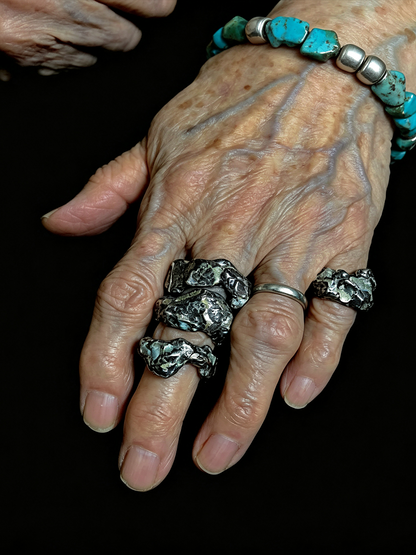 An elderly hand wearing multiple silver rings with textured, organic-shaped turquoise and dark stone inlays, alongside a matching turquoise beaded bracelet, set against a solid black background.