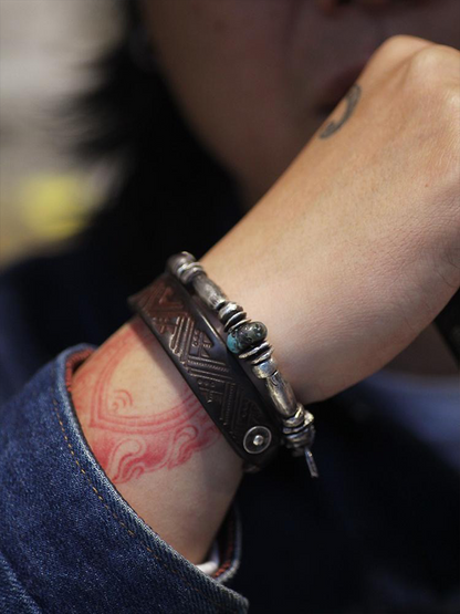 A close-up of a person's wrist wearing two bracelets: a dark engraved leather cuff bracelet and a silver-toned beaded bracelet with a small turquoise-like accent.