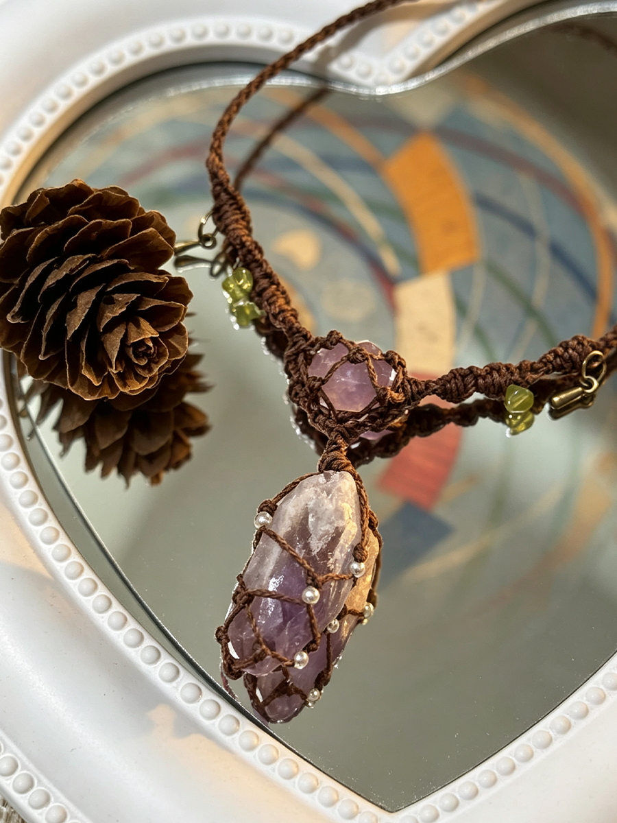 A handmade brown cord necklace featuring a large purple crystal pendant and smaller purple and green beads, displayed on a heart-shaped mirror with pinecones.